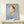 May include: Framed poster featuring a stylized cartoon girl with large green eyes, brown bob haircut, and yellow dress against a blue background with white geometric shapes. The poster displays "YOSHITOMO NARA" text and Japanese characters below. Shown in thin wooden frame on light wood surface next to small wrapped gift box with twine and greenery against cream wall.