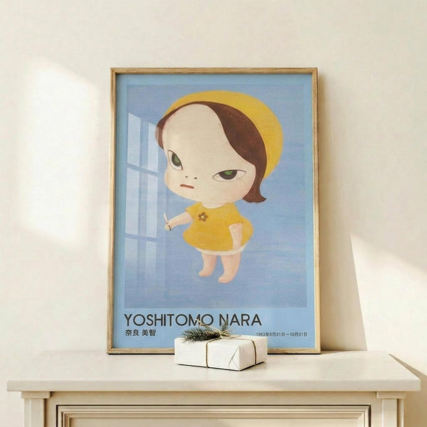 May include: Framed poster featuring a stylized cartoon girl with large green eyes, brown bob haircut, and yellow dress against a blue background with white geometric shapes. The poster displays "YOSHITOMO NARA" text and Japanese characters below. Shown in thin wooden frame on light wood surface next to small wrapped gift box with twine and greenery against cream wall.