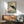 May include: A vintage-style illustration of a black crane with red crown standing among pink peonies and green foliage, displayed in a thin wooden frame above a mid-century modern wood credenza. The artwork features muted sage green and cream tones in an Asian-inspired botanical style. A small white ceramic table lamp sits on the credenza next to a gray upholstered armchair with wooden frame in a minimalist living room with white walls and natural sunlight casting shadows.