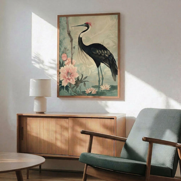 May include: A vintage-style illustration of a black crane with red crown standing among pink peonies and green foliage, displayed in a thin wooden frame above a mid-century modern wood credenza. The artwork features muted sage green and cream tones in an Asian-inspired botanical style. A small white ceramic table lamp sits on the credenza next to a gray upholstered armchair with wooden frame in a minimalist living room with white walls and natural sunlight casting shadows.