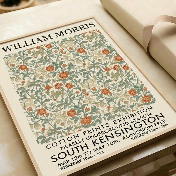 May include: Vintage-style exhibition poster featuring William Morris floral pattern in sage green and terracotta orange on cream background. The decorative botanical design shows intertwining vines and blooms. Text indicates cotton prints exhibition at South Kensington with dates and admission details. Poster appears unframed on white paper, photographed alongside rolled paper tubes on neutral surface.