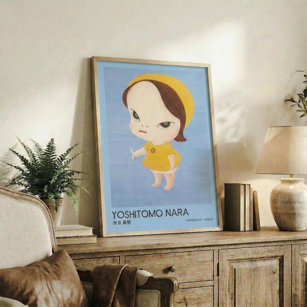May include: Framed poster featuring a stylized illustration of a young girl with brown hair, yellow beret and dress, holding a small knife, set against a soft blue background. The artwork is displayed in a light wood frame on a rustic wooden sideboard alongside books, pottery, and a fern plant in a cozy living room setting with beige furniture and neutral tones.