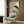 May include: Japanese-style artwork featuring a black crane with red head crest standing among pink peonies and water lilies, displayed in light wooden frame on wood-paneled wall above beige upholstered chair with geometric throw pillow, brass floor lamp and dark side table with open book nearby
