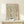May include: Framed vintage-style botanical poster featuring William Morris floral pattern in sage green, coral orange, and cream colors. The artwork shows intricate flowering vines and leaves in a repeating design, displayed in natural wood frame on light wooden shelf against white wall. Small wrapped gift box sits at base of frame.