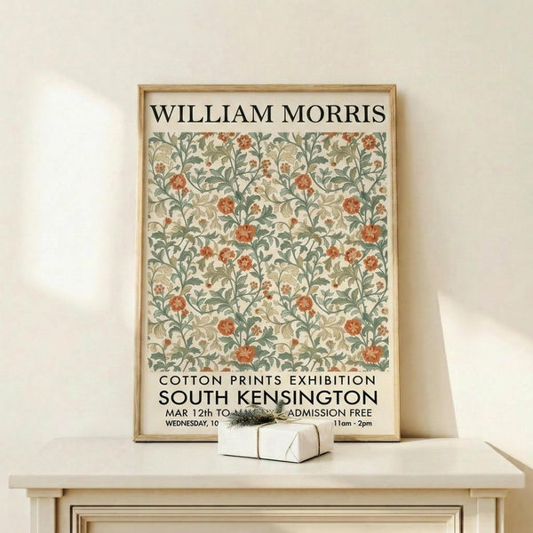 May include: Framed vintage-style botanical poster featuring William Morris floral pattern in sage green, coral orange, and cream colors. The artwork shows intricate flowering vines and leaves in a repeating design, displayed in natural wood frame on light wooden shelf against white wall. Small wrapped gift box sits at base of frame.