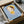 May include: Framed art print in black frame being unpacked from cardboard box with protective wrapping. The artwork shows a stylized cartoon character with short brown hair, yellow halo, green eyes, and yellow dress holding a small knife, set against a light blue background. Text at bottom reads "Yoshitomo Nara" with dates. Wooden frame corners and white tissue paper visible around the package.