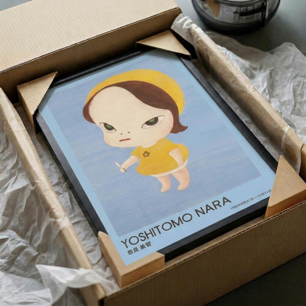 May include: Framed art print in black frame being unpacked from cardboard box with protective wrapping. The artwork shows a stylized cartoon character with short brown hair, yellow halo, green eyes, and yellow dress holding a small knife, set against a light blue background. Text at bottom reads "Yoshitomo Nara" with dates. Wooden frame corners and white tissue paper visible around the package.