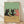 May include: Framed poster print leaning against white textured wall on light wood floor. Artwork shows three cats sitting at wooden table - two black cats and one orange cat - sharing wine with bottle and glass. Green background, cartoon illustration style in natural wood frame.