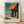 May include: Framed Edward Hopper poster print leaning against white textured wall on light wooden floor. The artwork shows a mid-century urban storefront scene with bright orange awning, dark green columns, and turquoise sky. Natural lighting creates soft shadows across the minimal room setting. Light wood frame surrounds the vintage-style art print with artist name displayed below the image.