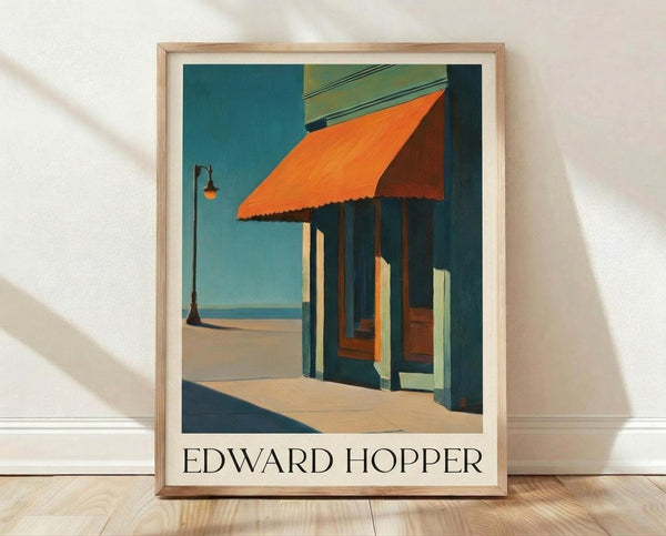 May include: Framed Edward Hopper poster print leaning against white textured wall on light wooden floor. The artwork shows a mid-century urban storefront scene with bright orange awning, dark green columns, and turquoise sky. Natural lighting creates soft shadows across the minimal room setting. Light wood frame surrounds the vintage-style art print with artist name displayed below the image.