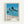 May include: Framed poster print hanging on white wall featuring stylized illustration of tall palm trees with long dark trunks against blue ocean water and beige sand beach. Clear blue sky background with geometric, clean art style. Light wood frame with white matting. Color palette dots and "TOKYO 1973" text at bottom in black lettering.