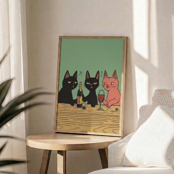 May include: Framed poster print featuring three cartoon cats at a wooden table - two black cats and one orange cat sharing wine with a bottle and glass, set against sage green background. Wood frame sits on round wooden side table next to beige upholstered chair with white pillow. Natural lighting creates soft shadows on cream wall with plant leaves visible in corner.