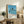 May include: Framed poster print displaying palm trees silhouetted against blue ocean and sky, styled as vintage travel poster with "Hiroshi Nagai Tokyo 1973" text and color dots at bottom. Light wood frame sits on rustic wooden sideboard alongside fern plant, ceramic vase, books, and table lamp in cozy living room setting with beige walls and leather armchair.