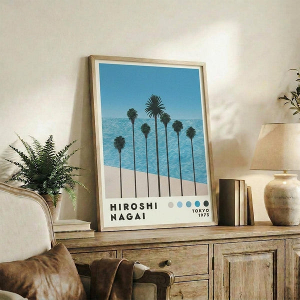 May include: Framed poster print displaying palm trees silhouetted against blue ocean and sky, styled as vintage travel poster with "Hiroshi Nagai Tokyo 1973" text and color dots at bottom. Light wood frame sits on rustic wooden sideboard alongside fern plant, ceramic vase, books, and table lamp in cozy living room setting with beige walls and leather armchair.