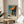 May include: Framed Edward Hopper poster print displayed on round wooden side table next to beige upholstered chair. Artwork shows architectural scene with orange awning, blue-green building facade, and desert landscape in mid-century American realist style. Natural sunlight creates shadows across white wall. Plant leaves visible in corner.