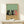 May include: Framed poster print showing three cartoon cats sitting at wooden table with wine bottle and glass. Two black cats and one pink/coral cat with sleepy expressions against sage green background. Natural wood frame displayed on white shelf next to small wrapped gift with greenery sprig.