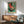May include: Vintage-style poster featuring a turntable with green and red color scheme and Japanese text, displayed in wooden frame above mid-century modern credenza. Room shows teal upholstered chair with wood arms, small white table lamp, and round coffee table. Natural sunlight creates shadows on white wall, giving the space a warm, retro aesthetic.