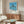 May include: Framed poster showing palm trees silhouetted against blue ocean and sky, displayed above a mid-century modern walnut credenza. White upholstered chair and oval glass coffee table visible in foreground. Poster features clean geometric design with beach scene and vintage typography. Room has neutral walls and natural lighting creating warm shadows on furniture.