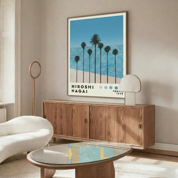 May include: Framed poster showing palm trees silhouetted against blue ocean and sky, displayed above a mid-century modern walnut credenza. White upholstered chair and oval glass coffee table visible in foreground. Poster features clean geometric design with beach scene and vintage typography. Room has neutral walls and natural lighting creating warm shadows on furniture.