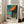 May include: Framed Edward Hopper poster leaning against white wall next to beige sofa in bright living room. Print shows architectural scene with orange awning over storefront columns, painted in Hopper's characteristic style with teal, orange and yellow tones. Light wood frame, approximately 2-3 feet tall, positioned on hardwood floor near large windows.