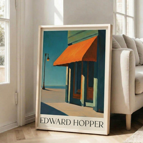 May include: Framed Edward Hopper poster leaning against white wall next to beige sofa in bright living room. Print shows architectural scene with orange awning over storefront columns, painted in Hopper's characteristic style with teal, orange and yellow tones. Light wood frame, approximately 2-3 feet tall, positioned on hardwood floor near large windows.