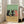 May include: Framed poster print leaning against white wall next to beige sofa in bright room with windows. Artwork shows three stylized cats sitting at wooden table - two black cats and one orange cat - sharing wine bottle and glass against sage green background. Light wooden frame, approximately 2-3 feet tall based on sofa scale.