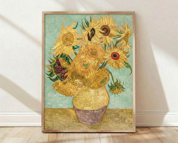 May include: Framed reproduction of Van Gogh's sunflowers painting showing vibrant yellow blooms with thick brushstrokes in a yellow vase against turquoise background, displayed in light wood frame leaning against white wall on wooden floor with natural lighting creating soft shadows