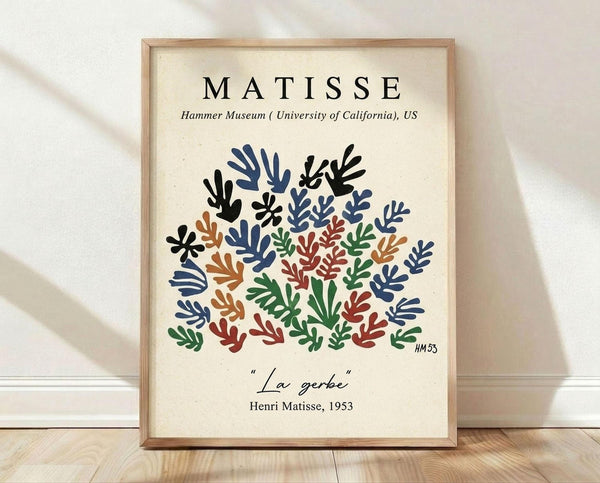 May include: Framed art print featuring colorful cut-out style botanical shapes in blue, green, red, orange and black arranged in a bouquet pattern. Text reads "MATISSE" at top, "Hammer Museum University of California US" below, and "La gerbe Henri Matisse, 1953" at bottom. Light wood frame against cream textured wall with white baseboard, sitting on light wooden floor with natural shadows.