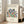 May include: Framed art print featuring colorful cut-out style botanical shapes in blue, green, orange, and red tones arranged in a circular pattern on cream background. Black text reads "MATISSE" at top and "La gerbe" at bottom. Light wood frame leaning against white wall next to beige upholstered chair in bright, airy room with large windows.