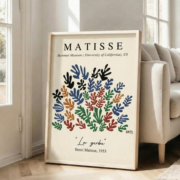 May include: Framed art print featuring colorful cut-out style botanical shapes in blue, green, orange, and red tones arranged in a circular pattern on cream background. Black text reads "MATISSE" at top and "La gerbe" at bottom. Light wood frame leaning against white wall next to beige upholstered chair in bright, airy room with large windows.