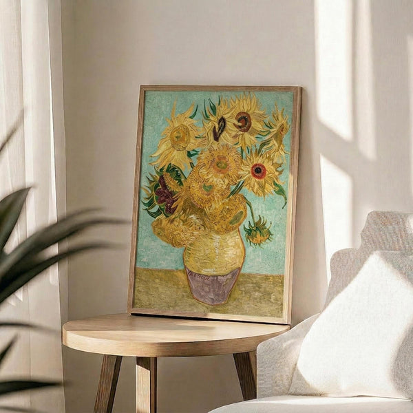 May include: Framed reproduction of Van Gogh's famous sunflowers painting displayed on round wooden side table next to beige upholstered chair. The artwork shows yellow sunflowers in a vase against turquoise background, housed in thin gold frame. Soft natural lighting creates shadows on cream wall, with plant leaves visible in corner of modern living room setting.