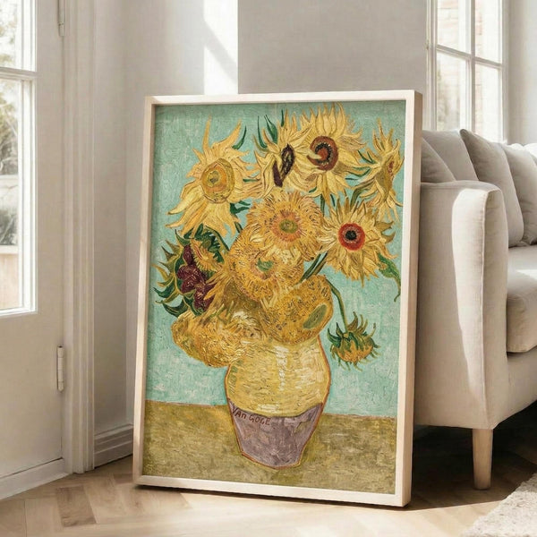 May include: Framed reproduction of Van Gogh's famous sunflowers painting leaning against white wall in bright living room. Yellow sunflowers in ceramic vase against turquoise background, displayed in light wood frame. Modern beige armchair and large windows with natural light visible in background. Artwork shows thick brushstrokes and vibrant colors characteristic of post-impressionist style.