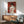 May include: Framed red and cream poster showing woman with upswept hair viewed from behind, wearing draped garment and holding roses, displayed above wooden sideboard with white table lamp in modern living room with mint green armchair and round coffee table, natural sunlight creating shadows on white wall
