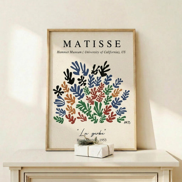 May include: Framed art print featuring Matisse's colorful cut-out style botanical design with blue, green, orange, and burgundy leaf shapes arranged in a circular pattern on cream background. Wooden frame sits on light wood shelf next to small wrapped gift with twine bow. Clean white wall backdrop creates minimalist home decor scene.