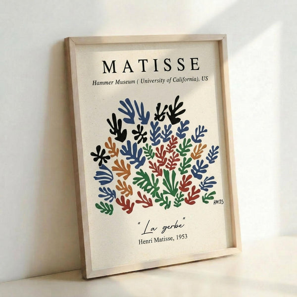 May include: Framed museum poster print featuring Matisse's colorful cut-out style botanical design with abstract leaf and coral shapes in blue, green, orange, red, and black on cream background. Light wood frame leaning against white wall, showing museum attribution text and artwork title below the vibrant organic pattern.