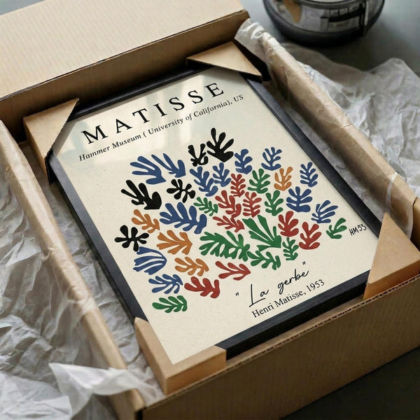 May include: Framed museum poster print featuring Henri Matisse's colorful botanical cut-out design with blue, red, green, orange and black leaf-like shapes arranged in circular pattern on cream background. Black frame sits in open cardboard shipping box with white tissue paper on wooden surface next to wrapped tube.