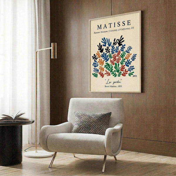 May include: Cream-colored poster with Henri Matisse's colorful botanical cutout design featuring blue, green, orange, and red leaf shapes arranged in a bouquet pattern. Shown framed in light wood frame hanging on wood-paneled wall above beige upholstered armchair with geometric throw pillow. Modern brass floor lamp and dark side table with open book visible in minimalist room setting.