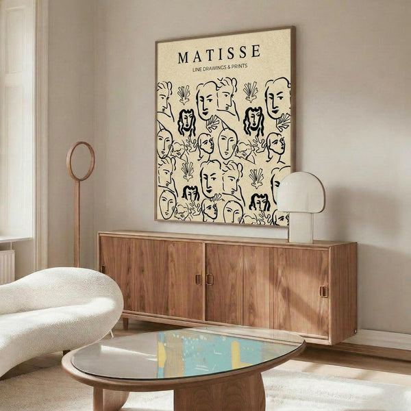 May include: Matisse-inspired poster with black line drawings of faces and botanical elements on cream background, displayed above mid-century wooden credenza with brass handles. Modern white curved chair and oval glass coffee table with colorful abstract art visible through glass top complete the minimalist living room setting with beige walls.
