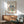 May include: Framed Matisse poster print hanging on white wall above wooden sideboard in modern living room. Print features colorful botanical cutout design with blue, green, red, and orange leaf shapes on cream background. Green upholstered mid-century chair with wooden arms sits beside sideboard. White ceramic table lamp and round wooden coffee table complete the scene. Natural sunlight creates shadows across the wall.