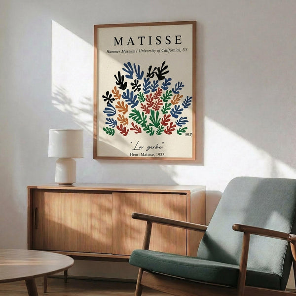 May include: Framed Matisse poster print hanging on white wall above wooden sideboard in modern living room. Print features colorful botanical cutout design with blue, green, red, and orange leaf shapes on cream background. Green upholstered mid-century chair with wooden arms sits beside sideboard. White ceramic table lamp and round wooden coffee table complete the scene. Natural sunlight creates shadows across the wall.