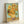 May include: Framed reproduction of Van Gogh's sunflowers painting leaning against white wall. Artwork shows vibrant yellow sunflowers in ceramic vase against turquoise background, displayed in light wood frame. Post-impressionist style with thick, textured brushstrokes visible throughout the composition.