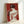 May include: Framed art print leaning against white wall showing woman from behind with dark hair in updo, holding red roses, wearing draped white garment with red linear details, set against deep red background in minimalist two-tone style, displayed in light wood frame on white surface