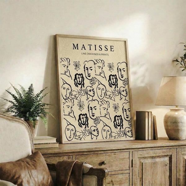 May include: Framed beige poster with black line drawings of faces and botanical elements in Matisse's minimalist style, displayed on rustic wooden sideboard alongside fern plant, ceramic vase, stacked books, and brown leather chair in neutral-toned living room setting.