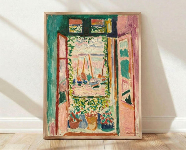 May include: Framed reproduction of Matisse's Fauve painting showing an interior view through open French doors to a harbor scene with boats. Vibrant greens, pinks, and oranges with bold brushstrokes. The artwork is displayed in a light wood frame against white walls on wooden flooring, with natural lighting casting soft shadows.