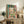 May include: Framed reproduction of Matisse's Fauve-style painting showing an open window with vibrant teal, coral, and pink colors, boats visible through the window, potted plants on the sill. The artwork leans against books on a rustic wood sideboard next to a fern plant, ceramic vase, and brown leather chair in a neutral-toned living room.