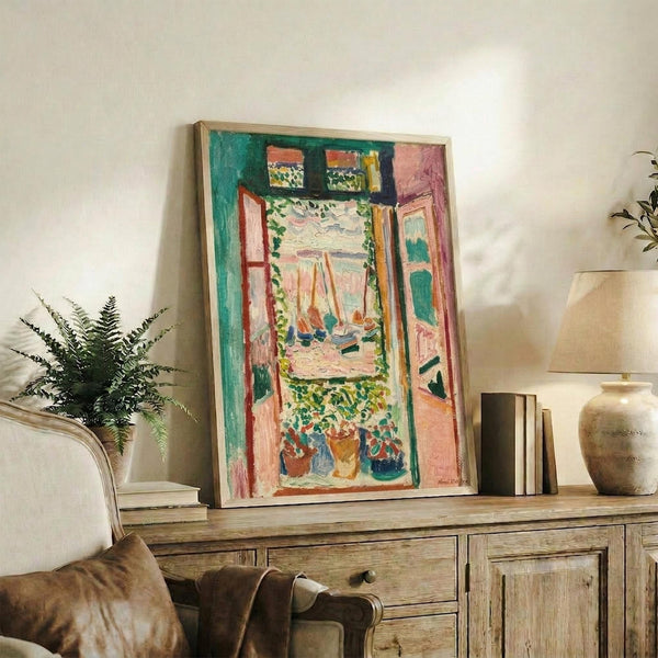 May include: Framed reproduction of Matisse's Fauve-style painting showing an open window with vibrant teal, coral, and pink colors, boats visible through the window, potted plants on the sill. The artwork leans against books on a rustic wood sideboard next to a fern plant, ceramic vase, and brown leather chair in a neutral-toned living room.