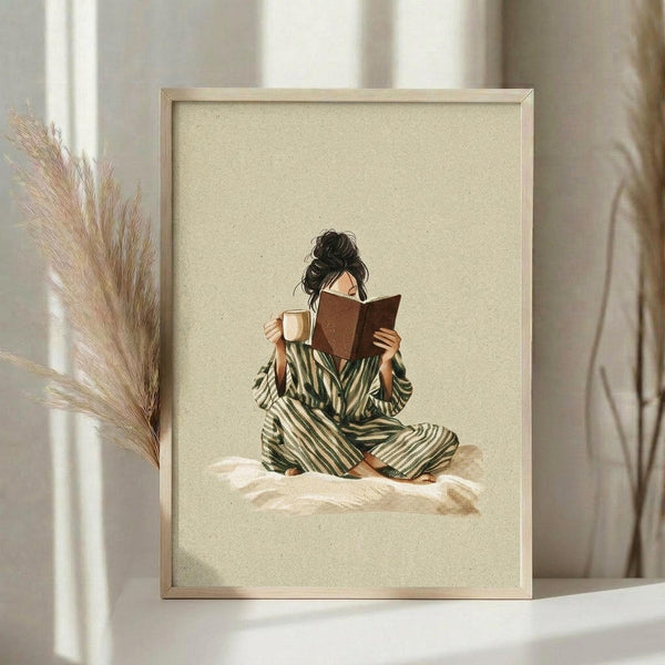 May include: Framed art print in light wood frame showing woman in green striped pajamas sitting cross-legged on bed reading book while holding coffee mug, with dark hair in messy bun, on cream textured background. Displayed on white surface next to dried pampas grass with soft natural lighting and neutral curtains in background.