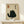 May include: Framed poster featuring a black cat illustration in Japanese ink painting style with yellow eyes, sitting pose, on cream textured background with black Japanese characters and red seal stamp. Natural wood frame displayed on light wooden shelf beside small wrapped white gift box with greenery sprig against white wall.