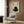 May include: Black cat artwork with Japanese calligraphy characters and red seal stamp on cream background, displayed in light wood frame above beige upholstered armchair with geometric patterned pillow. Modern reading nook with gold floor lamp, dark round side table with open book, and warm wood paneled walls.