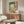 May include: Colorful Matisse-style painting depicting an open window with view of boats on water, hanging above a mid-century modern walnut credenza in a bright living room. The artwork shows vibrant greens, pinks, and blues in an expressionist style with loose brushstrokes. White modern chair and oval glass coffee table complete the contemporary interior setting with natural lighting.